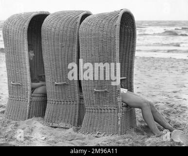 Drei große Basketballliegen an einem belgischen Strand, in denen zwei Personen sitzen. Sie wurden entwickelt, um maximalen Schatten vor der heißen Sonne zu bieten. Stockfoto