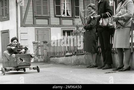 Ein Junge fährt mit seinem Gokart, beobachtet von drei Erwachsenen, die aussehen, als würden sie auf einen Bus warten. Stockfoto