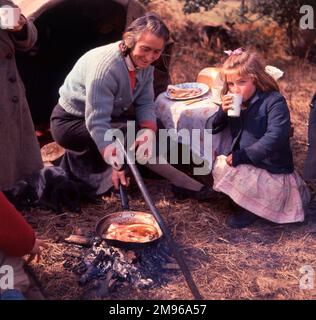 Eine Zigeunerin, die im Freien ein gebratenes Frühstück kocht. Ein kleines Mädchen hockt in der Nähe und schlürft eine Tasse Tee. Stockfoto