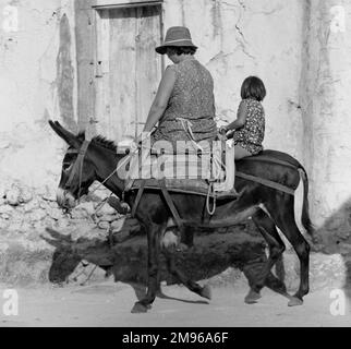 Eine ziemlich dicke Frau und ein kleines Mädchen, das an einem sonnigen Tag auf einem kleinen Esel reitet. Stockfoto