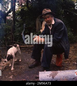 Ein Zigeuner und sein Hund, in einem Lager in Surrey. Der Mann sitzt unsicher auf einer zerknitterten roten Dose. Stockfoto