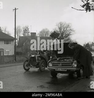 Ein kaputtes modernes Auto wird von einem Veteranenmodell in einer Vorstadtstraße überholt. Ein Mann schaut sich den Motor seines Limousinenwagens an, während ein paar in edwardianischem Kostüm vorbeifahren und vielleicht am London-Brighton-Lauf teilnehmen. Glücklicherweise ist der Mann Mitglied der AA, also sollte Hilfe bald auf dem Weg sein! Stockfoto