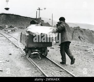 Ein Arbeiter, der eine Schieferbahnbahn schiebt, auf der ein großer Schieferblock ruht, irgendwo in Nordwales. Diese kleinen Straßenbahnen können bis zu einer halben Tonne Rohschiefer transportieren. Stockfoto