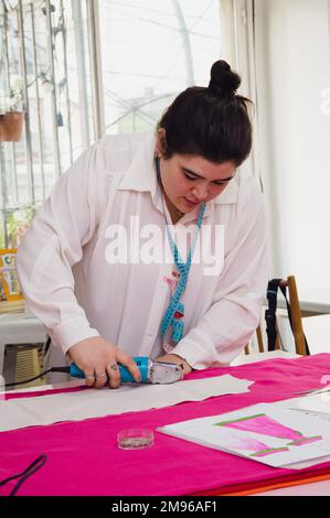 Vertikales Bild einer Kaukasierin, einer Modedesignerin in ihrer Werkstatt, die mit einem elektronischen Stoffschneider Gewebe schneidet Stockfoto