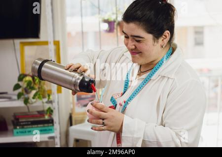 Junge weiße argentinierin, argentinische Ethnie, Modedesignerin, stehend, lächelnd, schüttet Wasser in ihre Partnerin, bereitet sich auf die Arbeit in h vor Stockfoto