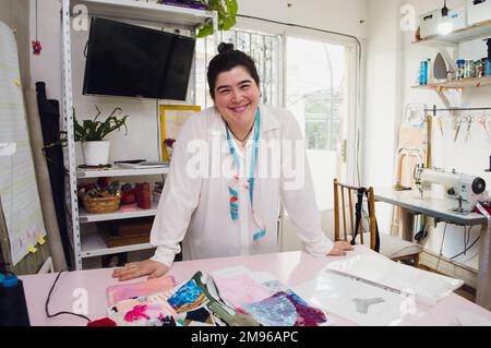 Porträt einer jungen Kaukasierin, die in ihrem Workshop für Kleidung zu Hause fröhlich lächelt, auf der Werkbank steht und in die Kamera schaut Stockfoto