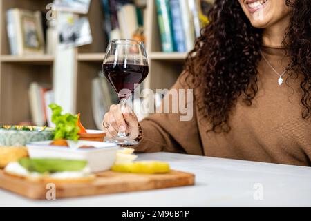 Nahaufnahme eines Glases Rotwein, das eine lächelnde junge Frau in der Hand hält, während sie eine Auswahl an Gemüse und veganen Speisen isst Stockfoto