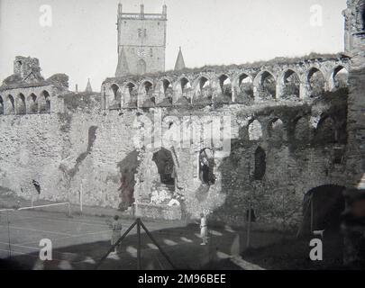 Ein Tennisplatz, auf dem Menschen spielen, in den Ruinen des mittelalterlichen Bischofspalastes, St. David's, Pembrokeshire, Dyfed, Südwales. Eine Reihe von Bögen im gotischen Stil, in denen früher Fenster zu sehen waren, mit dem Turm der Kathedrale in der Nähe. Einige Teile des Gebäudes stammen aus dem 12. Jahrhundert, aber die meisten Arbeiten wurden im 14. Jahrhundert ausgeführt. Stockfoto