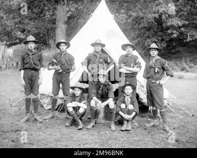 Eine Gruppe von acht Pfadfindern posiert für ihr Foto auf einem Feld, vor einem Zelt. Einer von ihnen hält ein Horn in der Hand, das sie zweifellos morgens alle aufgeweckt hat. Stockfoto