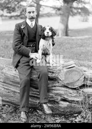 Ein Mann mittleren Alters sitzt auf einem großen Baumstamm in einem Landhausgarten irgendwo in Mid Wales, mit einem Spaniel an seiner Seite. Stockfoto