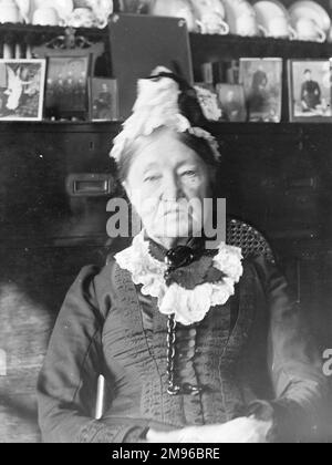 Eine ältere Frau in viktorianischem Kostüm, die vor einer walisischen Kommode sitzt, Mid Wales. Die Kommode enthält Familienfotos und Geschirr. Stockfoto
