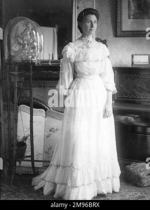 Eine elegante Dame in einem durchgehenden weißen Kleid posiert in einem Landhaus in Mid Wales. Es gibt einen Kamin und dekorierten Bildschirm auf der linken Seite und ein aufrechtes Klavier auf der rechten Seite. Stockfoto