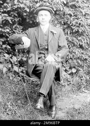 Ein schicker junger Mann sitzt auf einem Klappstuhl in einem Garten, Mid Wales. Er trägt einen dreiteiligen Anzug mit einer Blume im Knopfloch und einer flachen Mütze. Stockfoto