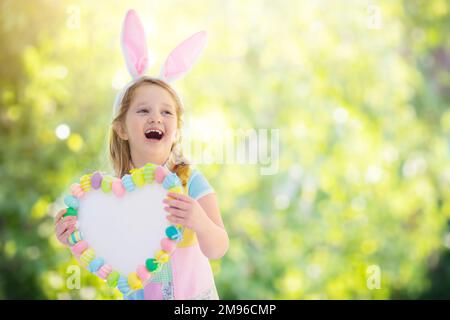 Glückliches kleines Mädchen mit Hasenohren, das ein Herzbrett mit bunten Ostereiern hält. Kinder feiern Ostern. Kinder haben Spaß auf der Ostereiersuche. Stockfoto