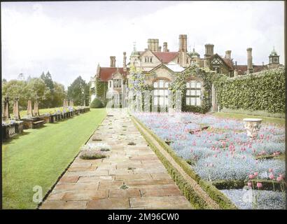 Blick auf den Garten und das Haus in Paddockhurst, nahe Selsfield Common, West Sussex. Das Haus stammt aus dem 17. Jahrhundert und wird heute als Schule genutzt. Hier sehen Sie einen breiten Pfad mit Gras auf der linken Seite (einschließlich einer Steinbalustrade) und einem langen Blumenrand auf der rechten Seite. Ivy wächst entlang der Gartenwand und auf einem Teil des Hauses. Stockfoto