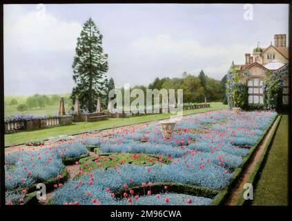Blick auf den Garten und das Haus in Paddockhurst, nahe Selsfield Common, West Sussex. Das Haus stammt aus dem 17. Jahrhundert und wird heute als Schule genutzt. Hier sehen Sie einen langen Blumenrand mit Blumen und Sträuchern, mit zwei Ziervasen. Ivy wächst auf einem Teil des Hauses. Auf der linken Seite ist eine Steinbalustrade mit Blick über einige Felder. Stockfoto