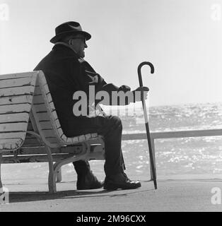 Ein alter Mann mit einem Gehstock, der auf einer Bank mit Blick aufs Meer sitzt. Stockfoto