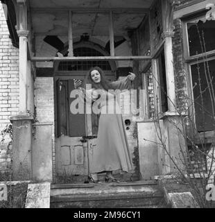 Eine junge Frau in einem langen Kleid posiert künstlerisch vor der Tür eines heruntergekommenen Hauses mit kaputten Fenstern. Stockfoto