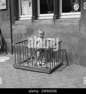 Ein kleiner Junge steht in einem Laufstall auf einem Gehweg vor einem Café. Stockfoto