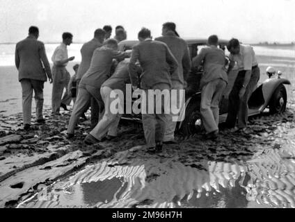 Eine Gruppe von Männern, die ein Auto auf einen nassen Sandstrand schieben. Stockfoto