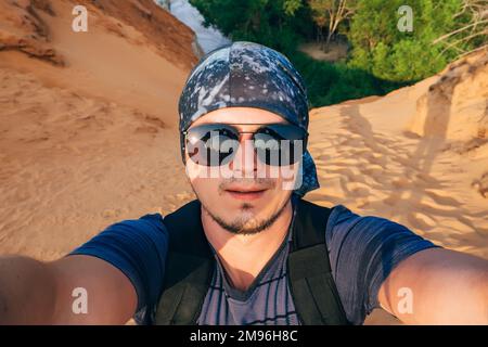 Man wandert mit einem Rucksack und macht unterwegs ein Selfie auf dem Kamerafläch in einer Sonnenbrille und einem Bandana Stockfoto