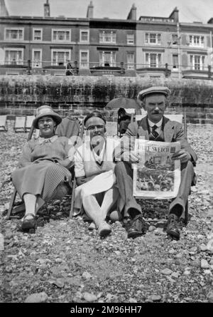 Drei Urlauber, zwei Frauen und ein Mann, sitzen am Kieselstrand in Hastings, Sussex. Er liest die Zeitung Daily Sketch. Eine Hotelterrasse ist im Hintergrund zu sehen. Stockfoto