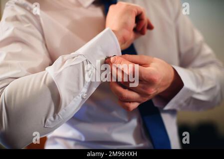 Der Bräutigam legt am Hochzeitstag Manschettenknöpfe auf den Ärmeln eines weißen Hemds. Die Hände eines Mannes mit einer Krawatte Stockfoto