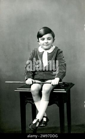 Der kleine Junge sitzt auf dem Hocker, der Stock in der Hand, posiert für die Kamera Stockfoto