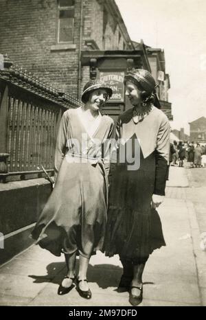 Zwei Frauen, in langen Kleidern und Glockenhüten, gehen zusammen eine Straße entlang. Stockfoto