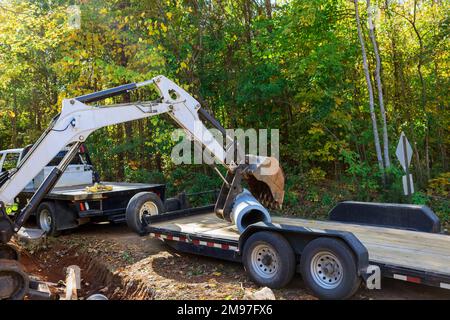 Da Betonkläranlagen für unterirdische Installationen vorbereitet werden, verwendet der Arbeiter den Traktor, um das Rohr in den Boden zu heben. Stockfoto