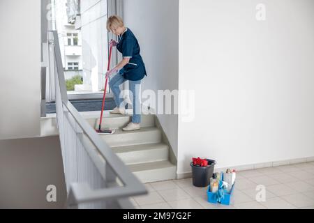 Die Putzfrau in Uniform mit Mopp und Lappen wäscht staubige Stufen des Hauses Stockfoto