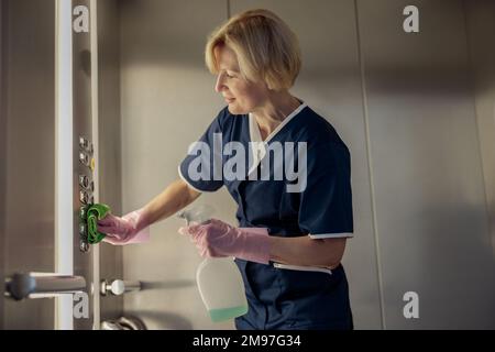 Professionelles Zimmermädchen trägt Uniform und Handschuhe, reinigt den Aufzug mit Reinigungsmittel und Lappen Stockfoto