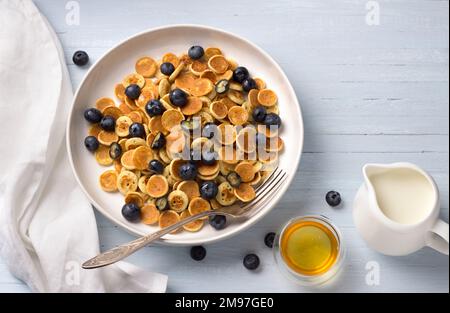 Teller mit Mini-Pfannkuchen, Müsli mit Sahne, Honig und Beeren auf hellblauem Hintergrund. Trendiges Essen. Hausgemachtes Frühstück. Draufsicht. Leerzeichen Stockfoto