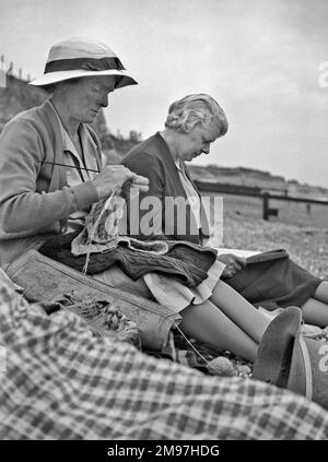 Zwei Frauen mittleren Alters, die an einem Kieselstrand sitzen, eine strickend, die andere liest. Stockfoto