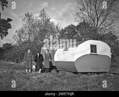 Drei Leute und ein Hund auf einem Feld, neben zwei Wohnwagen. Stockfoto