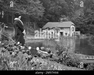 Eine Frau, die an einem See steht, mit Tulpen im Vordergrund und einer strohgedeckten Hütte in der Ferne. Stockfoto