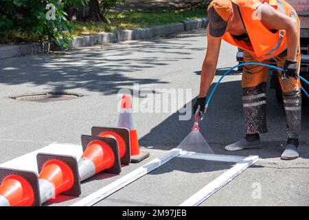 Ein Straßenarbeiter bringt an einem Sommertag weiße Straßenmarkierungen auf einer Fahrbahn mit einer Airbrush an. Speicherplatz kopieren. Stockfoto