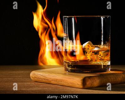Glas mit Whiskey mit Eis auf Holztisch und schwarzem Hintergrund mit Feuer Stockfoto