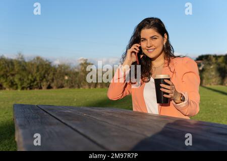 Eine lateinische Geschäftsfrau, die an einem Holztisch in einem Park sitzt und am Telefon telefoniert und einen Kaffee hält. Stockfoto