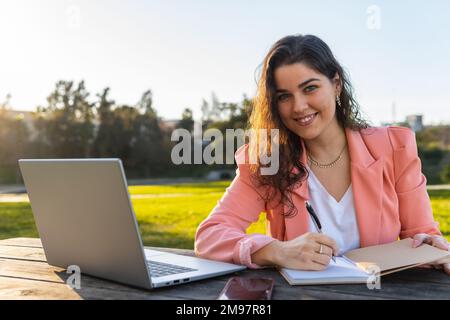 Eine Geschäftsfrau, die lächelnd in die Kamera schaut und ihre Notizen in ihr Notizbuch schreibt, während ihr Computer neben ihr in einem Park im Freien steht. Stockfoto