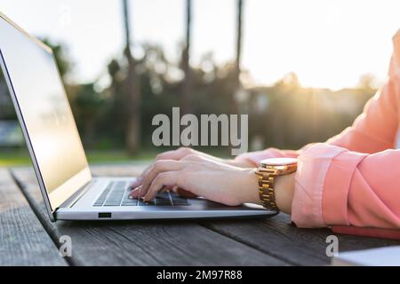 Unbekannte Person an einem Holztisch in einem Park, die ihren Laptop im Freien benutzt. Stockfoto