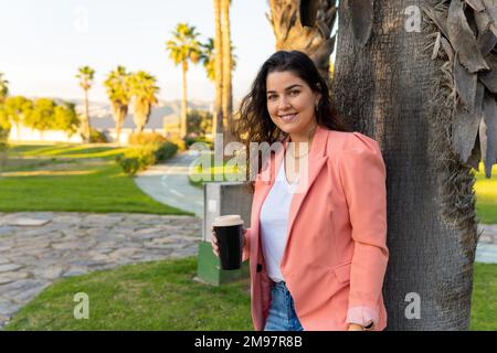 Gut gekleidetes, junges Latina-Mädchen, das ihr Glas Kaffee hält und sich in einem Park an einen Baum lehnt. Stockfoto