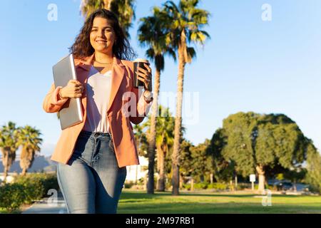 Gut gekleidete, junge Geschäftsfrau aus Latina, die in der Kamera lächelt und ihren Laptop und Kaffee in der Hand hält. Stockfoto