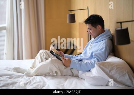 Ein hübscher, junger asiatischer Mann, der auf dem Bett eine Zeitschrift las. Seitenansicht Stockfoto