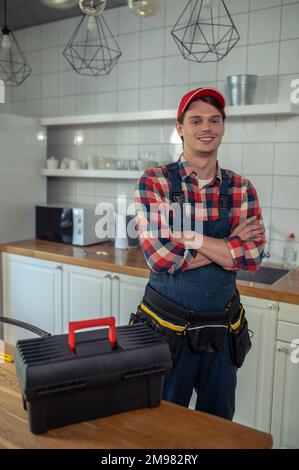 Lächelnder, glücklicher Mechaniker in einem Overall, der neben dem Werkzeugkasten stand, der auf dem Küchentisch platziert wurde Stockfoto