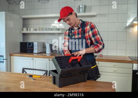 Seriöser junger weißer Handwerker in Arbeitskleidung, der den Werkzeugkasten auf dem hölzernen Küchentisch öffnet Stockfoto