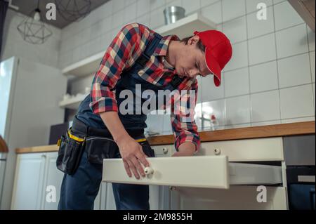 Ruhiger, fokussierter Mechaniker in Arbeitskleidung, der den Knopf an der Vorderseite der Schublade des Küchenschranks befestigt Stockfoto