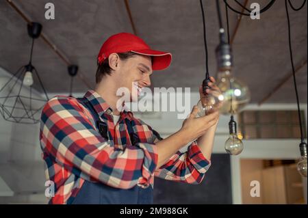 Porträt eines fröhlichen Elektrikers in der Kappe, der die Glühlampe in die Fassung schraubt Stockfoto