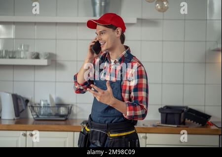 Lächelnd zufriedener junger, kaukasischer Reparaturtechniker, der in der Kundenküche mit dem Mobiltelefon sprach Stockfoto