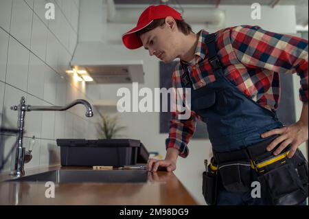 Ernsthaft fokussierter junger weißer Klempner in der Arbeitskleidung, der den Wasserhahn in der Kundenküche überprüft Stockfoto